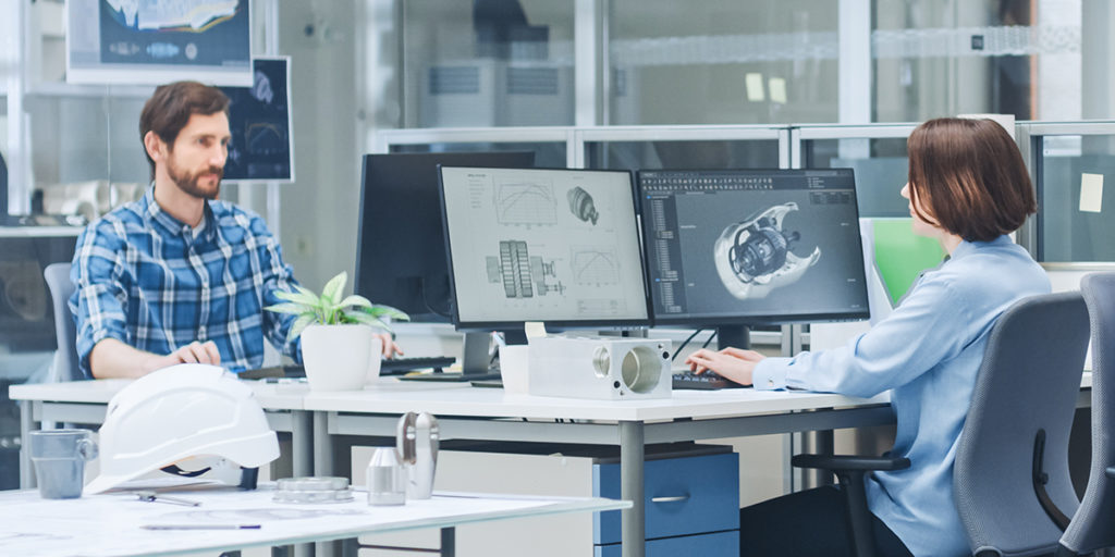 two people sitting at desks in an office with computer monitors between them